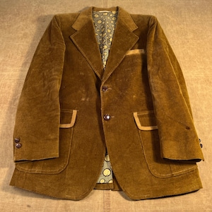 May include: A brown corduroy blazer with a notched lapel, two front pockets, and a breast pocket. The blazer has a two-button closure and brown buttons. The interior lining features a patterned design. The blazer is a vintage style.