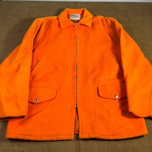 Peut inclure: Veste orange vif avec une fermeture éclair intégrale, deux poches avant avec fermetures à boutons argentés et un col. La veste a un aspect doux et texturé et une étiquette au col.