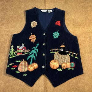 Puede incluir: Chaleco de pana azul marino con bordados de temática otoñal. Presenta calabazas, hojas, un granero y un gato negro. El chaleco tiene cuello en V, cierre de botones y un corte entallado. Una prenda festiva para el otoño.