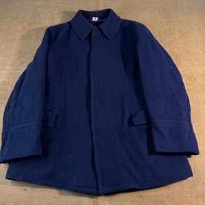 May include: A dark navy blue wool-blend coat featuring a classic fold-down collar, long sleeves with wide cuffs, and two front flap pockets. The garment has a concealed front closure and a small pink rectangular tag at the inner neckline.