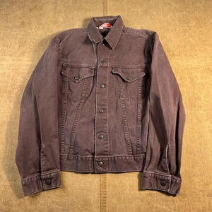 May include: A dark brown denim jacket featuring a classic collar, a full-length front button closure, and two chest pockets with buttoned flaps. The jacket has long sleeves with buttoned cuffs and visible stitching details throughout the garment.