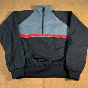 May include: A black and gray anorak jacket with a red stripe across the chest. The jacket has a stand-up collar, a half-zip closure, and elastic cuffs and hem. The jacket is made of a lightweight, water-resistant material.