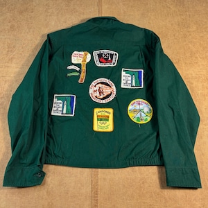 May include: A dark green long-sleeve jacket featuring several embroidered patches on the back. The patches include text such as Blackhawk District, Wah-Lo-Ha Trail, Frankfort Kentucky, Camporee, and She-Ka-Gong Trail, along with various graphic illustrations.