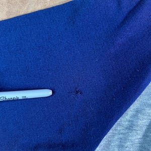May include: Close-up of a blue and gray baseball style shirt sleeve. A white Sharpie fine point pen is resting on the blue fabric. The shirt has a raglan sleeve design.