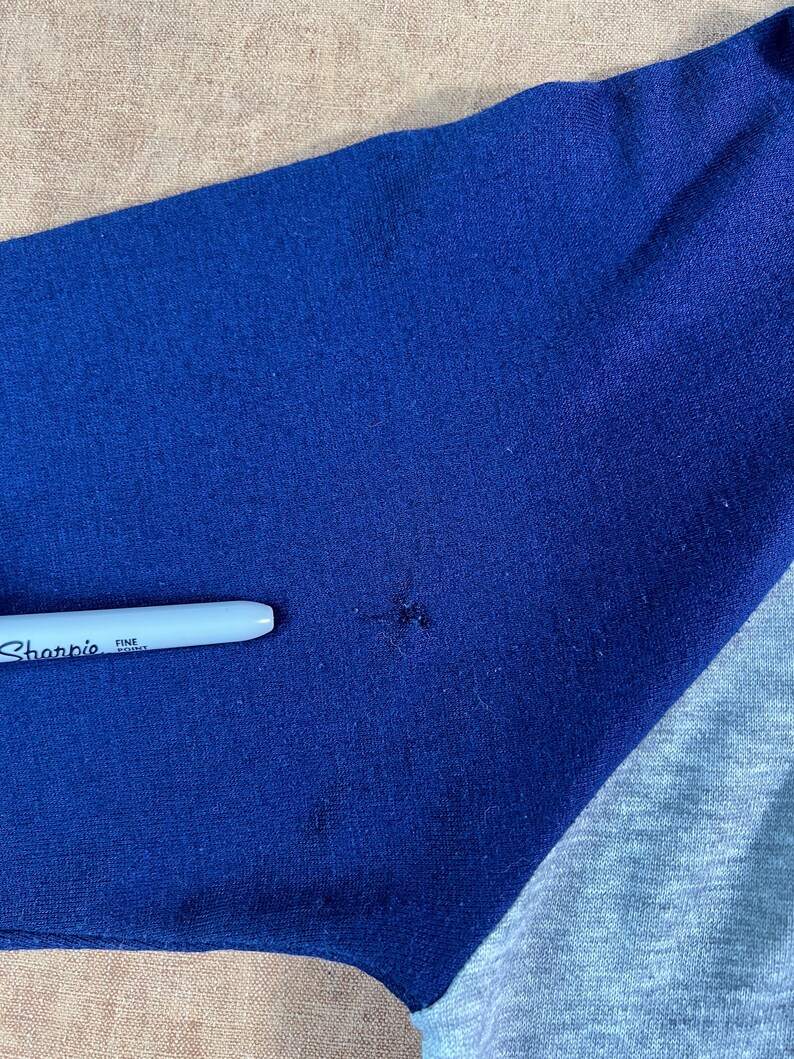 May include: Close-up of a blue and gray baseball style shirt sleeve. A white Sharpie fine point pen is resting on the blue fabric. The shirt has a raglan sleeve design.