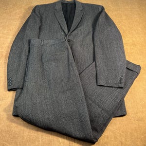 Puede incluir: Un traje gris carbón, que consta de una chaqueta y un pantalón a juego. La chaqueta tiene solapas con muescas y cierre de botón. Los pantalones son de frente plano. El traje está hecho de una tela texturizada.