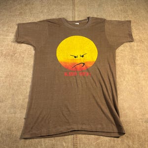 May include: Brown t-shirt with a yellow and orange angry face graphic. The graphic shows a frowning mouth and angled eyebrows. The text "KISS OFF" is printed in red below the face. The t-shirt has short sleeves and a crew neck.