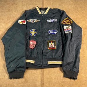 May include: A dark navy blue zip-up bomber jacket featuring various embroidered automotive brand patches including Dodge, Chevrolet, Ford, Chrysler, and Nash. The jacket has a ribbed collar and hem with white and gold stripes and long sleeves.