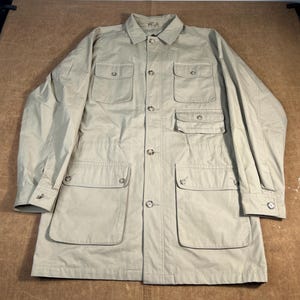 May include: A tan-colored long jacket with multiple pockets and a button-up front. The jacket has a collared neckline and buttoned cuffs. The jacket is made of a lightweight material.