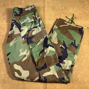 Pode incluir: Calças cargo camufladas com um padrão de floresta em tons de verde, castanho e preto. As calças têm vários bolsos, fechos de botão e cordões nos tornozelos. O tecido parece ser um material durável de estilo militar.