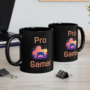 Puede incluir: Dos tazas de cerámica negras con un diseño blanco. El diseño presenta un mando de videojuego con un fondo de puesta de sol y el texto "Pro Gamer".