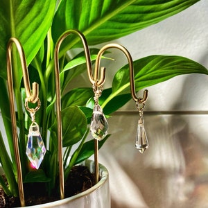 Op de afbeelding: Drie gouden metalen standaards met kristallen traanvormige bedels die eraan hangen. De standaards staan in een witte en roze keramische pot met een groene plant.