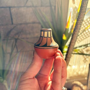 May include: A small, rounded terracotta-colored ceramic vessel featuring a narrow neck and a wide, bulbous base. The upper portion is decorated with a series of vertical black lines and geometric shapes that contrast against the natural clay surface.