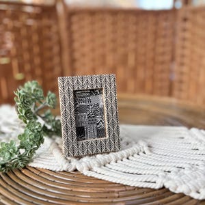 May include: A small, rectangular picture frame with a black and white geometric pattern. The frame holds a black and white patterned image. The frame is displayed on a white macrame table runner.