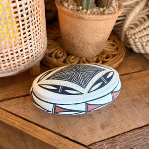 May include: An oval-shaped ceramic box with a white base, featuring black geometric line patterns on the lid and a series of black and terracotta-colored triangular motifs around the side of the container, creating a distinct decorative appearance.