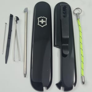 Könnte beinhalten: Ein schwarzes Victorinox Schweizer Taschenmesser-Griffset mit silbernem Wappenlogo, schwarzem Taschenclip und Zubehör wie einer Metallnadel, einem schwarzen Kunststoffwerkzeug, einer silbernen Pinzette, einem Metallstift und einem neongelben Schlüsselband.