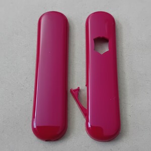 Könnte beinhalten: Zwei längliche Kunststoffobjekte in Magenta. Eines hat einen sternförmigen Ausschnitt und einen kleinen, angebrachten Arm. Das andere ist ein solider, glatter und abgerundeter Rechteck. Beide haben eine glänzende Oberfläche.