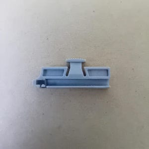 Könnte beinhalten: Ein hellgrauer Plastikclip mit einem erhöhten Mittelteil und zwei kleineren Abschnitten auf jeder Seite. Der Clip ist dazu bestimmt, Gegenstände zusammenzuhalten.