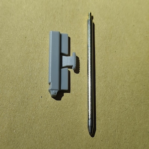 Puede incluir: Un pestillo de plástico gris con un pasador de metal. El pestillo tiene una pequeña perilla texturizada en el lateral. El pasador mide aproximadamente 15 cm de largo y tiene una punta puntiaguda.