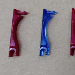 Op de afbeelding: Vier plastic onderdelen in verschillende kleuren: transparant, rood, blauw en roze. Elk onderdeel is gebogen en heeft aan één uiteinde een haak.