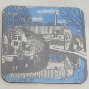May include: A blue and grey illustrated coaster featuring a quaint village scene with a church steeple, a bridge, and a reflection of the village in the water.
