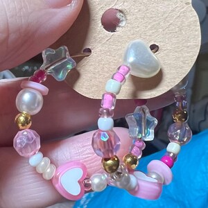 May include: Two pink and white beaded hoop earrings with heart and star charms. The earrings are made with small pink, white, and iridescent beads.