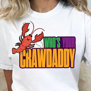 K&ouml;nnte beinhalten: Wei&szlig;es T-Shirt mit einem roten Krebs, der eine Kette aus gr&uuml;nen und violetten Perlen tr&auml;gt. Der Text "Who's Your Crawdaddy" ist in Gelb, Gr&uuml;n und Lila gedruckt.