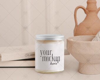 Puede incluir: Un tarro de vela blanco con una tapa plateada y una etiqueta que dice "your mockup here". La vela está sobre una mesa blanca con una superficie de madera marrón debajo. Hay un jarrón de cerámica marrón y un manojo de salvia en el fondo.