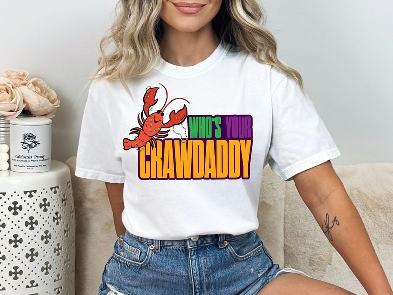 K&ouml;nnte beinhalten: Wei&szlig;es T-Shirt mit einem roten Krebs, der eine Kette aus gr&uuml;nen und violetten Perlen tr&auml;gt. Der Text "Who's Your Crawdaddy" ist in Gelb, Gr&uuml;n und Lila gedruckt.