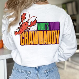 K&ouml;nnte beinhalten: Wei&szlig;es Langarm-T-Shirt mit einem Cartoon-Krebs, der Mardi Gras-Perlen tr&auml;gt und den Text "Who's Your Crawdaddy" in Lila, Gr&uuml;n und Gelb.