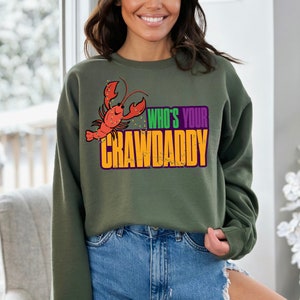 K&ouml;nnte beinhalten: Gr&uuml;nes Sweatshirt mit einem Cartoon-Krebs und dem Text "Who's Your Crawdaddy" in Orange, Lila und Gr&uuml;n.