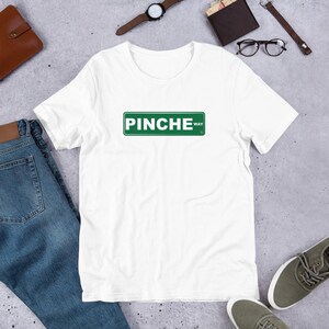 May include: White t-shirt with a green and white street sign graphic that reads "PINCHE WAY". The shirt is short-sleeved and appears to be made of a soft material. The shirt is laying on a light gray surface.