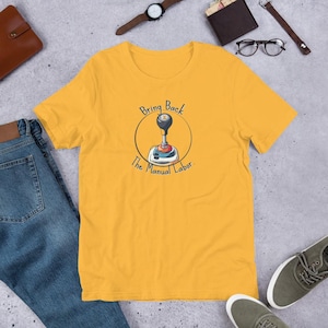 May include: Mustard yellow t-shirt featuring a graphic of a car's gear shift with the text "Bring Back The Manual Labor." The design is in a circle. The shirt is made of a soft material.