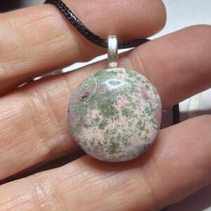 May include: A round, polished pendant featuring a mottled pattern of sage green and soft pink mineral inclusions. The stone is set in a simple silver-toned metal bail and is suspended from a thin, black braided cord necklace.