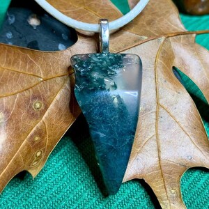 May include: A handcrafted pendant featuring a polished, triangular gemstone with a silver bail. The stone displays a blend of deep teal and translucent hues, with a white cord. The pendant is displayed on a brown leaf.