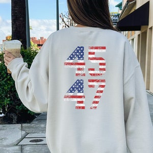 May include: A light beige sweatshirt with a distressed American flag design featuring the numbers "45 47" printed on the back. The sweatshirt is made of a soft, comfortable material.