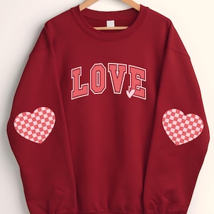 May include: Red sweatshirt with the word "LOVE" in white letters with a pink heart above it. The sweatshirt has pink and white checkered hearts on the sleeves.