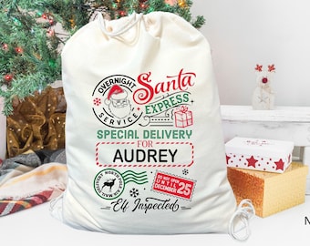 May include: A white canvas Santa sack with red and green text that reads "Overnight Santa Express Service - Special Delivery for Audrey - Elf Inspected". The sack has a drawstring closure and is decorated with a Santa Claus illustration, a reindeer, and a gift box.
