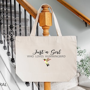 May include: A natural canvas tote bag with the text "Just a Girl Who Loves Hummingbird" and a colorful hummingbird graphic.