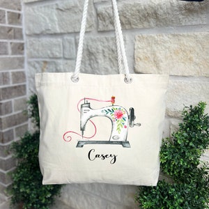 May include: Cream-colored tote bag with white rope handles. The bag features a watercolor illustration of a sewing machine adorned with flowers and the name "Casey" in black script. The sewing machine has pink thread.