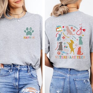 May include: A gray t-shirt with a paw print and the name "HANNAH" on the front. The back of the shirt has a colorful design with a variety of veterinary-related icons, including a stethoscope, a thermometer, a syringe, and a paw print. The text "VETERINARY TECH" is printed in a colorful font on the back of the shirt.