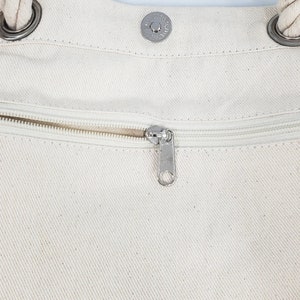 May include: Close-up of a cream-colored canvas tote bag. The bag features a silver zipper, a magnetic snap closure, and rope handles attached with silver rings. The bag's texture is visible, and the zipper is partially open.