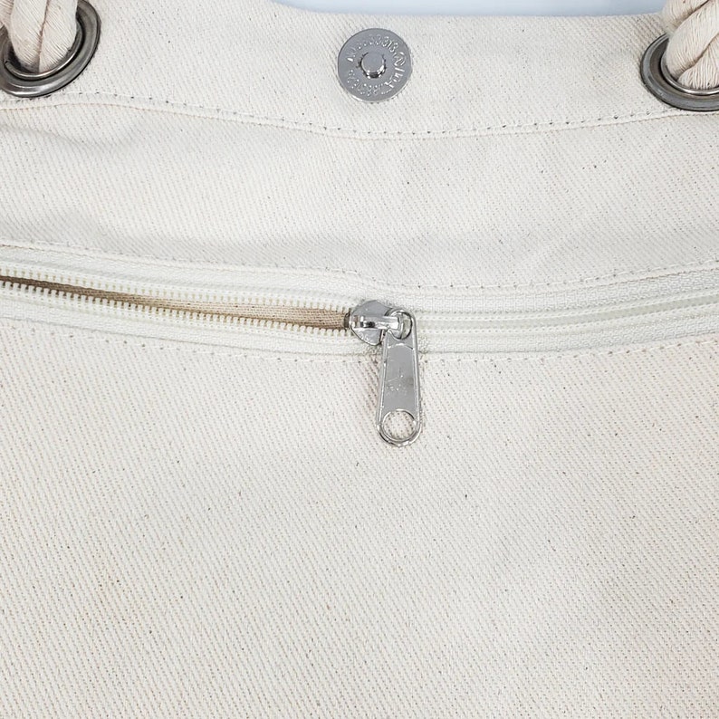 May include: Close-up of a cream-colored canvas tote bag. The bag features a silver zipper, a magnetic snap closure, and rope handles attached with silver rings. The bag's texture is visible, and the zipper is partially open.