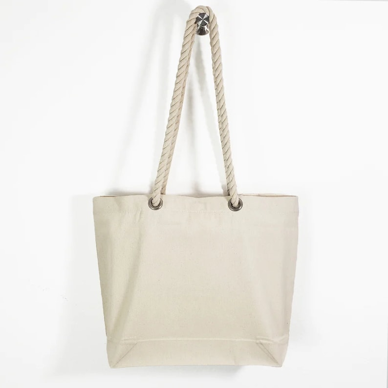 May include: A cream-colored canvas tote bag with rope handles. The bag has a rectangular shape with a flat bottom and is suspended from a silver hook. The rope handles are thick and beige, attached to the bag with silver grommets.