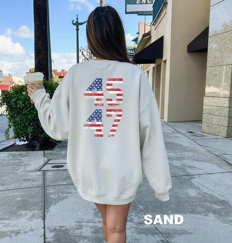May include: A light beige sweatshirt with a distressed American flag design featuring the numbers "45 47" printed on the back. The sweatshirt is made of a soft, comfortable material.