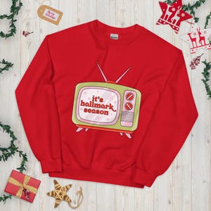 May include: A red sweatshirt with a retro television graphic. The television is green and pink, with two red knobs and antennas. The text "it's hallmark season" is in pink and red. A festive holiday item, perfect for the Christmas season.