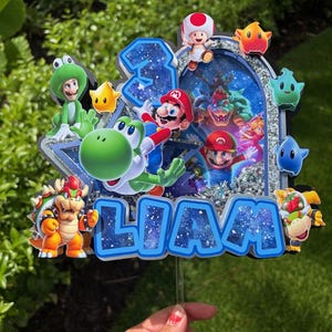 K&ouml;nnte beinhalten: Ein mehrschichtiger Tortenaufleger im Super Mario-Design mit dem Namen LIAM in blauen Buchstaben mit Galaxie-Muster, der Zahl 3 und verschiedenen Charakter-Ausschnitten wie Mario, Yoshi, Bowser und Toad vor einem silbernen Glitzerrahmen.