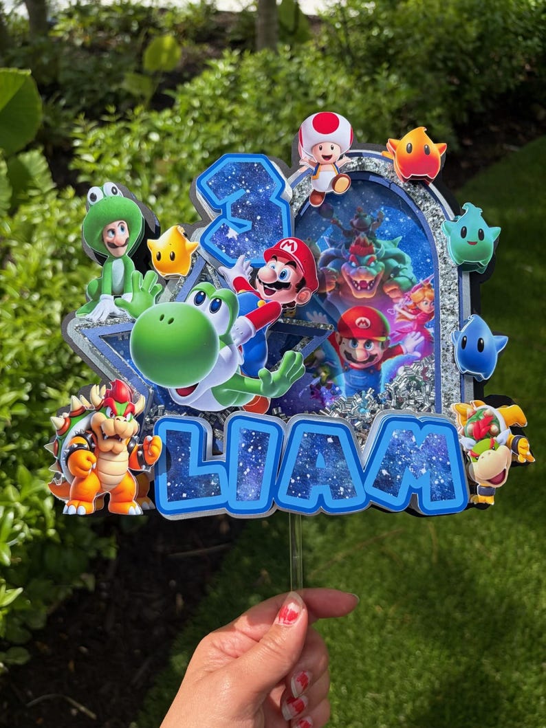 K&ouml;nnte beinhalten: Ein mehrschichtiger Tortenaufleger im Super Mario-Design mit dem Namen LIAM in blauen Buchstaben mit Galaxie-Muster, der Zahl 3 und verschiedenen Charakter-Ausschnitten wie Mario, Yoshi, Bowser und Toad vor einem silbernen Glitzerrahmen.