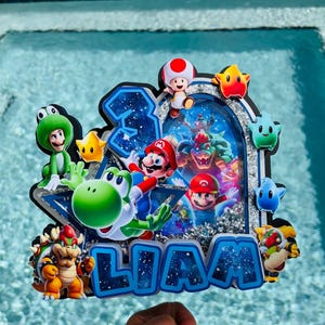 K&ouml;nnte beinhalten: Ein individueller Tortenaufleger mit mehrschichtigem Design, der die Zahl 3 und den Namen LIAM in blauer Weltraum-Optik zeigt. Er enth&auml;lt bunte Ausschnitte von Mario, Yoshi, Bowser, Toad und verschiedenen Sternen vor einem silbernen Glitzerhintergrund.