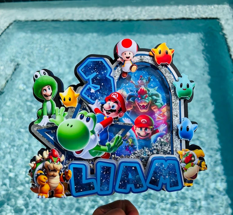 K&ouml;nnte beinhalten: Ein individueller Tortenaufleger mit mehrschichtigem Design, der die Zahl 3 und den Namen LIAM in blauer Weltraum-Optik zeigt. Er enth&auml;lt bunte Ausschnitte von Mario, Yoshi, Bowser, Toad und verschiedenen Sternen vor einem silbernen Glitzerhintergrund.